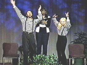 A woman holds a book and two mimes in striped shirts leap up with their hands raised.