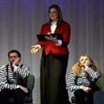 A woman holds a book, and gestures to two mimes in striped shirts.