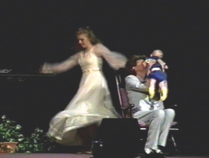A dancer in white flowing dress, dances beside a woman, on a rocking chair, holding a baby.